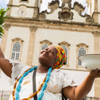 A woman dressed in traditional clothes from Bahia, holding ingredients to cook.
